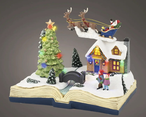 Miniatyrjulscen på en öppen bok med snötäckt hus, barn, bro, belyst julgran och jultomten i sin släde.
