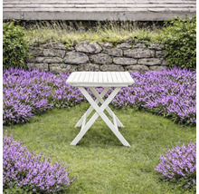 Vitt hopfällbart trädgårdsbord i trä med ribbad yta på en gräsmatta omgiven av lila blommor och en stenmur.