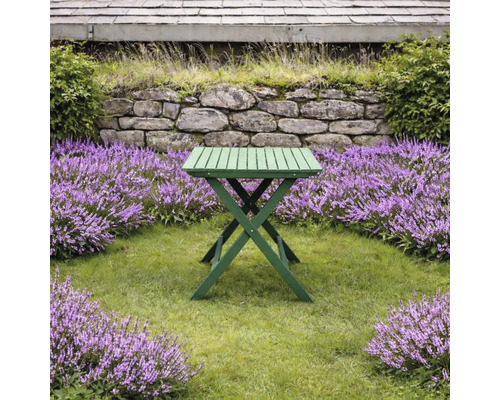 Grönt fällbart träbord i en trädgård med gräsmatta, lila blommor och stenmur.