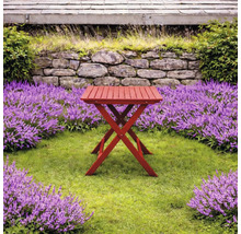 Rött hopfällbart sidobord i trä i en ljus trädgård med gräsmatta, lila blommor och en stenmur.