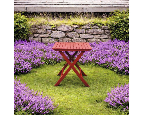 Rött hopfällbart sidobord i trä i en ljus trädgård med gräsmatta, lila blommor och en stenmur.