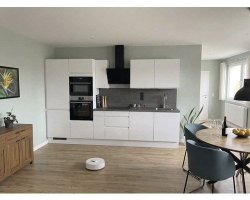 Modernt kök med vita skåp, grått stänkskydd, runt träbord, blå stolar och robotdammsugare på ljust trägolv.