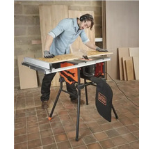 Man med hörselskydd och handskar använder en Black and Decker bordscirkelsåg med stativ för att kapa en träskiva.