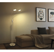 Modern golvlampa i metall med två justerbara huvuden i ett vardagsrum bredvid en soffa med kuddar och tavlor.