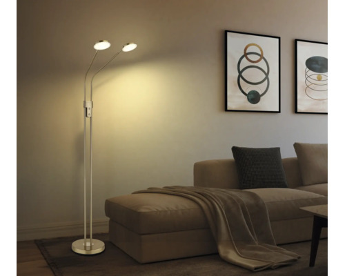 Modern golvlampa i metall med två justerbara huvuden i ett vardagsrum bredvid en soffa med kuddar och tavlor.