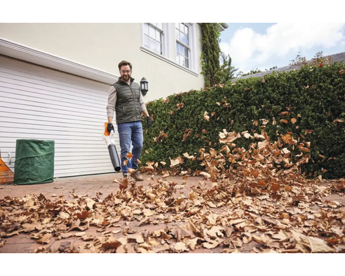 En man använder en lövblås från STIHL för att ta bort torra löv från en stenlagd garageuppfart framför ett hus med garage och häck.