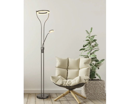 Modern golvlampa med ringformad uplight och läsarm bredvid en fåtölj och en krukväxt i ett ljust rum.
