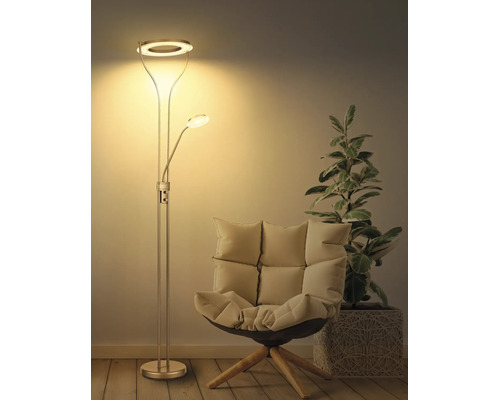 Modern golvlampa i metall med uplight och läsarm i ett vardagsrum bredvid en fåtölj och en växt. Varm belysning.