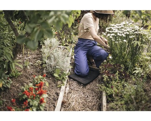 En person knäböjer på ett knäskydd vid trädgårdsarbete i en rabatt med olika blommande växter och buskar.