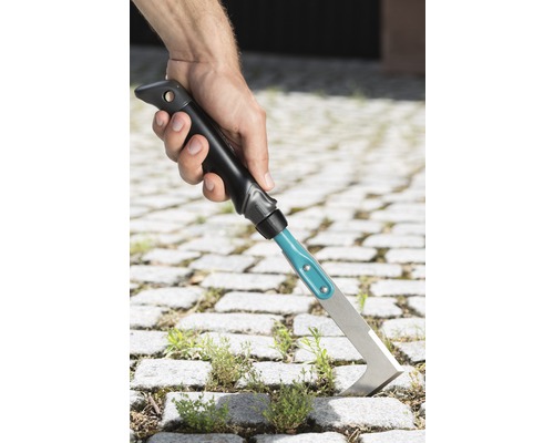 Hand håller en fogskrapa för att ta bort ogräs mellan gatstenar.