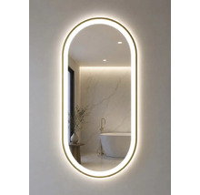 Oval badrumsspegel med guldram och LED-belysning. Spegeln reflekterar ett ljust badrum med fristående badkar och marmorvägg.
