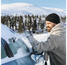 En man med mössa och stickad tröja tar bort snö från bilens vindruta med en isskrapa i ett snöigt vinterlandskap.