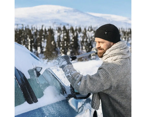 En man med mössa och stickad tröja tar bort snö från bilens vindruta med en isskrapa i ett snöigt vinterlandskap.
