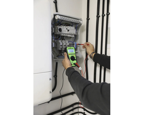 Elektriker mäter spänning i en öppen säkringslåda med en digital KPS-multimeter för elektrisk feldiagnostik.