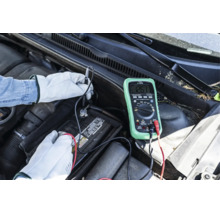 Person med handskar mäter spänningen på ett bilbatteri med en digital multimeter i motorutrymmet.