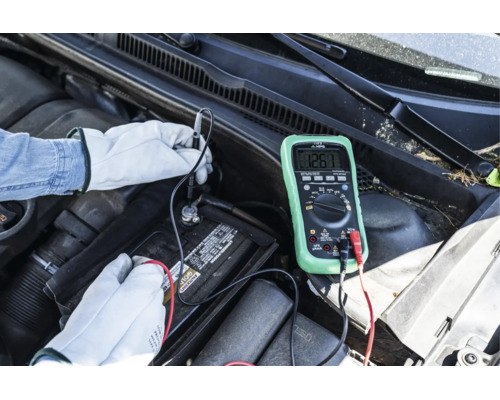 Person med handskar mäter spänningen på ett bilbatteri med en digital multimeter i motorutrymmet.