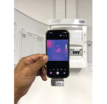 En hand håller en smartphone med en värmekamera framför ett öppet säkringsskåp för kontroll av elanläggningen.