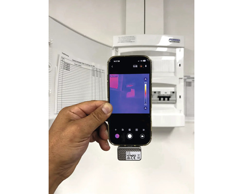 En hand håller en smartphone med en värmekamera framför ett öppet säkringsskåp för kontroll av elanläggningen.