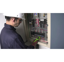 En elektriker med skyddshjälm kontrollerar ledningarna i ett öppet elskåp med en spänningsprovare.