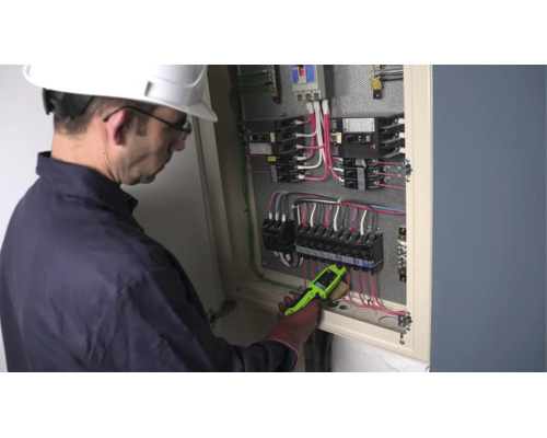 En elektriker med skyddshjälm kontrollerar ledningarna i ett öppet elskåp med en spänningsprovare.