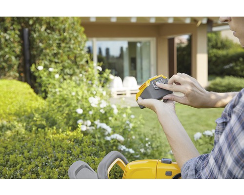 En person trycker på knappen på ett gult batteri för en häcksax i trädgården.