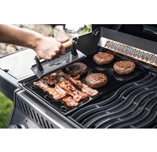 På en grill ligger hamburgare och bacon som bearbetas med en grillpress.