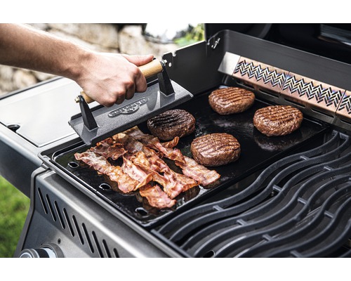 På en grill ligger hamburgare och bacon som bearbetas med en grillpress.