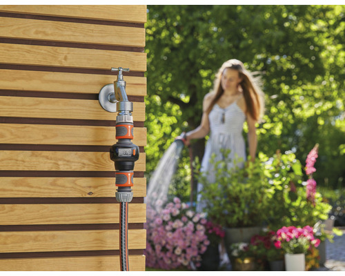 Trädgårdsvattenmätare på vattenkran med kvinna i bakgrunden som vattnar trädgården