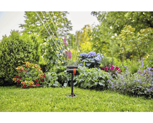 Trädgårdssprinkler i drift på gräsmatta med blomsterbäddar i bakgrunden.