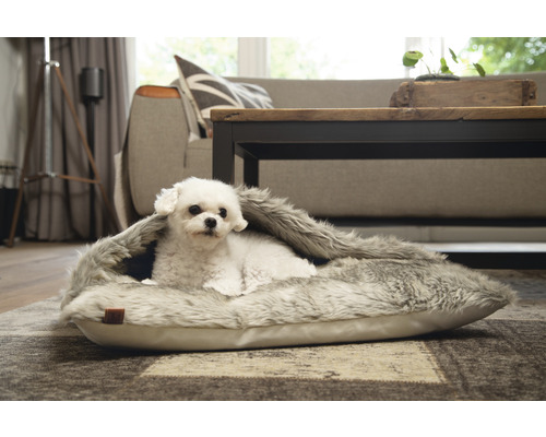 En vit hund ligger i en grå djurbädd i ett vardagsrum.