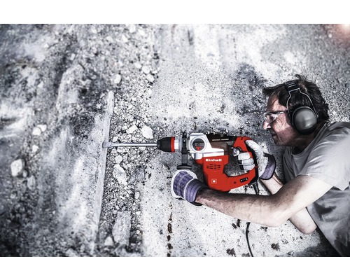 En man borrar i en betongvägg med en borrhammare. Borrhammaren är röd och silverfärgad och kommer från Einhell.