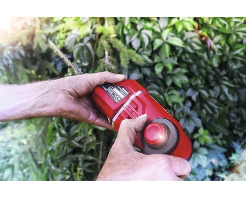 Person använder en sladdlös grässax framför en grön häck.