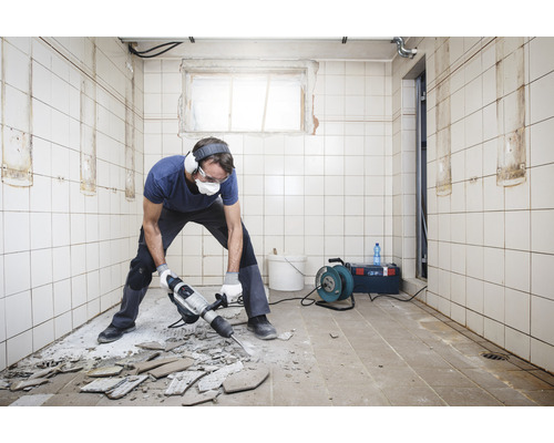 Man med skyddsglasögon och hörselskydd slår bort kakel från ett golv med en borrhammare