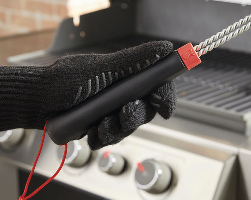 En hand i en grillvante håller en grillborste framför en grill.