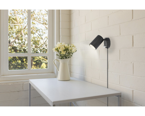 Modern scen med en svart vägglampa, ett vitt bord och en vas med blommor bredvid ett fönster.