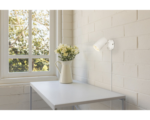 Miljöbild med ett vitt konsolbord, en vas med blommor och en vägglampa