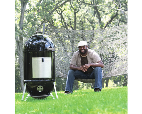 Vertikal kolgrill bredvid en person i en hängmatta i trädgården.