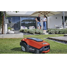 En robotgräsklippare står på en gräsmatta framför ett hus med ett par i bakgrunden.