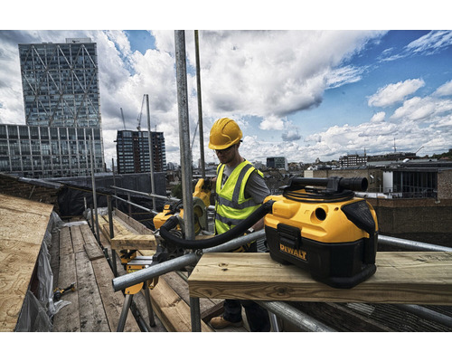 Hantverkare med skyddshjälm och varselväst arbetar med en våt- och torrdammsugare från Dewalt på en byggnadsställning framför en stadsbild.