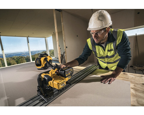 En byggnadsarbetare med hjälm och skyddsglasögon skär en skiva med en DeWalt gipsskiva i ett grovt rum. En DeWalt dammsugare står i bakgrunden.