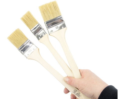 En hand håller tre penslar med trähandtag och naturliga borst