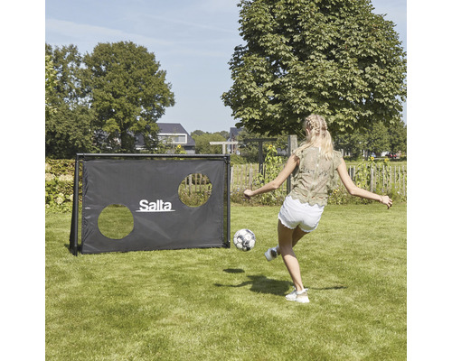 En kvinna sparkar en fotboll mot en fotbollsmålvakt i trädgården. Målet har två hål och en Salta logotyp.