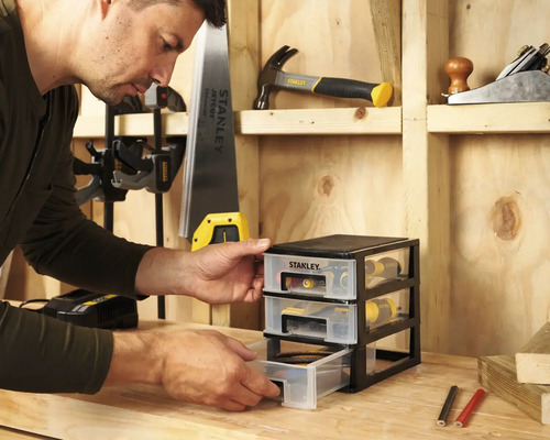 Man öppnar en låda i en Stanley verktygsorganisatör på en arbetsbänk framför en trävägg med verktyg.