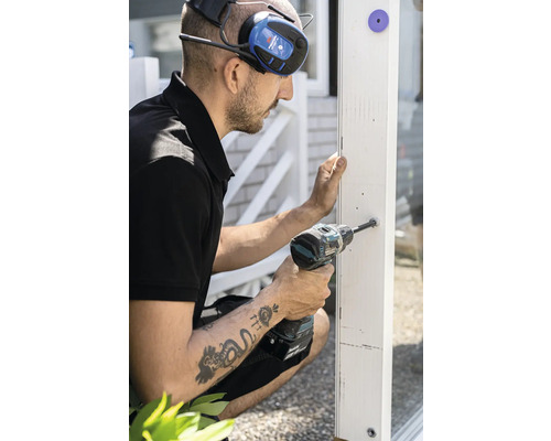 Hantverkare borrar ett hål i en vit träkomponent med en sladdlös skruvdragare. Han bär hörselskydd.