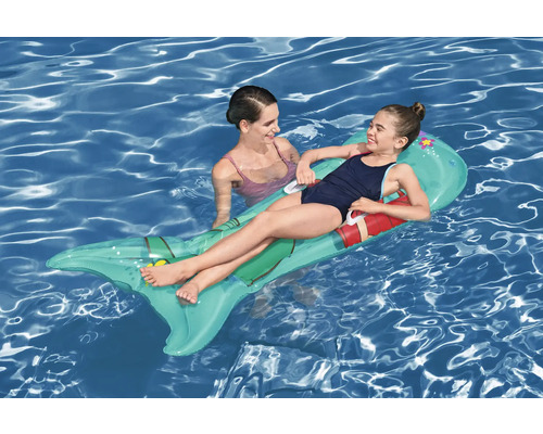 Flicka ligger på en uppblåsbar fisk luftmadrass i vattnet, bredvid henne står en kvinna.