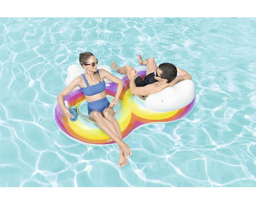 Två personer kopplar av på en uppblåsbar regnbågs-poolö i vattnet.