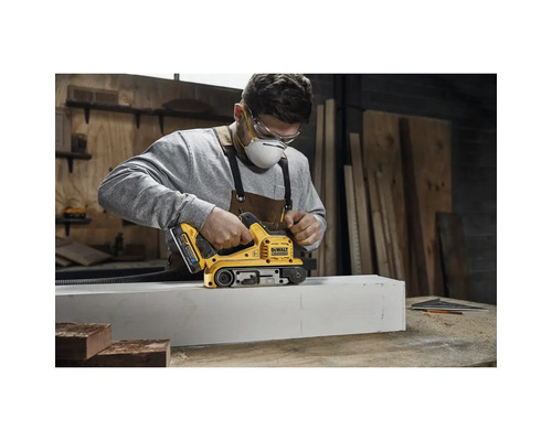 En man använder en bandslip från DeWalt för att bearbeta ett arbetsstycke. Han bär ett skyddsmask och skyddsglasögon.