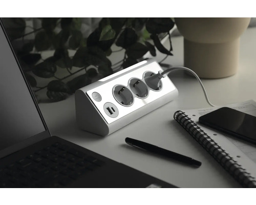 Grenuttag med tre jordade uttag och två USB-portar på ett skrivbord med bärbar dator, penna, anteckningsbok och smarttelefon.