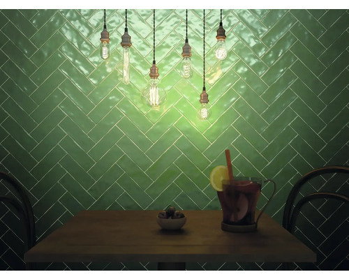 Väggdekoration med gröna plattor i fiskbensmönster, belyst med glödlampor ovanför ett bord med drycker