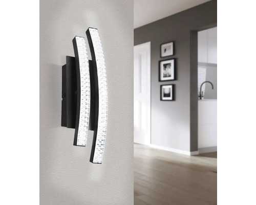 Modern vägglampa med två böjda, upplysta element och ett mörkt fäste i ett ljust rum.